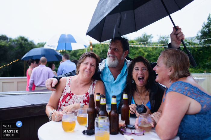 El fotógrafo de bodas de Outer Banks capturó esta foto de invitados de bodas disfrutando de la recepción al aire libre a pesar de la lluvia, ya que todos comparten un paraguas