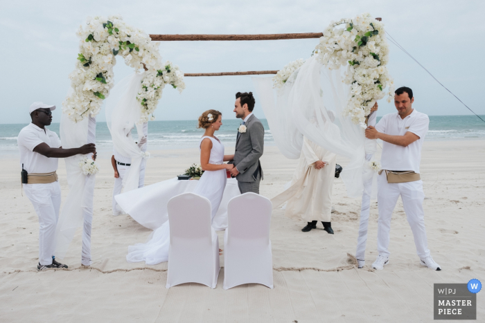 Fotograf ślubny z Dubaju robi zdjęcie prywatnej ceremonii na plaży, podczas gdy pomocnicy pomagają utrzymać łuk ślubny pod wiatr