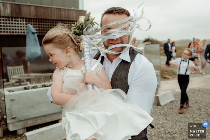 Bristol wedding photographer captured this photo of a flower girls boquet ribbons being blown into the grooms face as he carries her across a parking lot