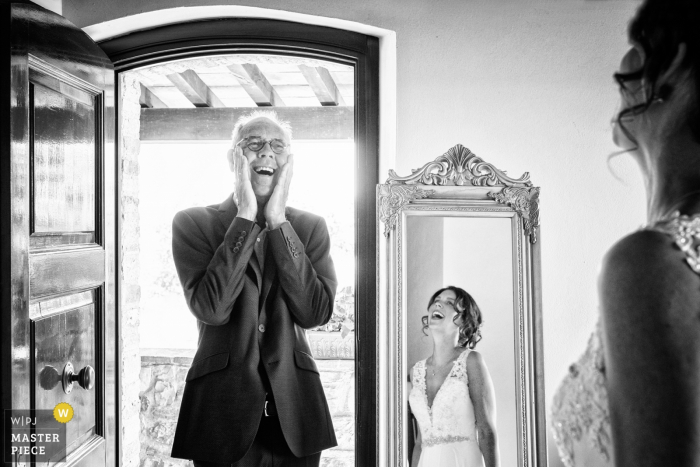 El fotógrafo de bodas de Arezzo capturó esta foto en blanco y negro de la encantadora expresión del padre de las novias al ver a su hija con su vestido de novia por primera vez. La podemos ver riendo en el espejo frente a ella en la habitación.