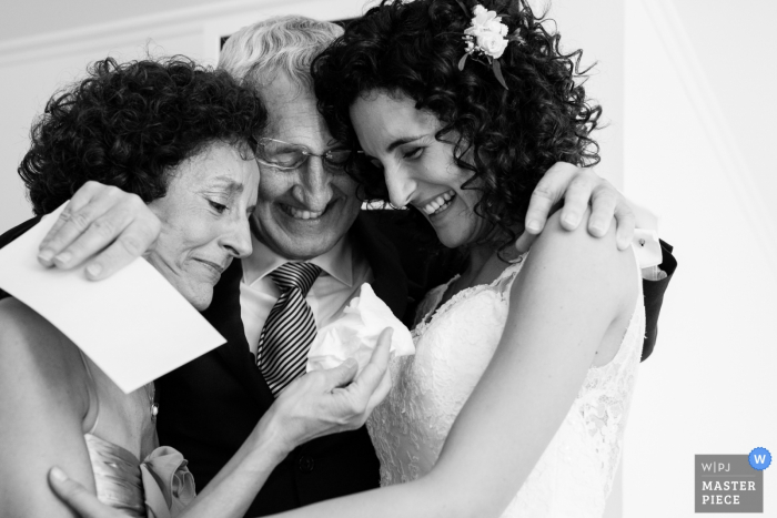 Il fotografo di matrimoni di Barcellona ha catturato questa foto in bianco e nero di una sposa sorridente che abbraccia entrambi i genitori mentre aspettano che inizi la cerimonia