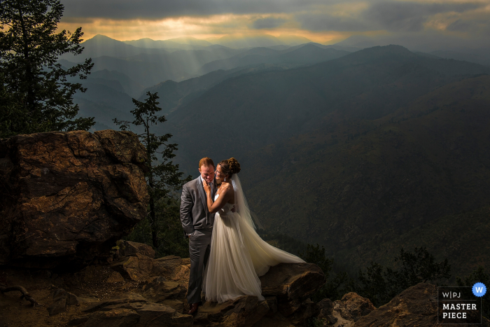 Photo by: <a href="https://www.wpja.com/contests/2017-tri3-photography-competition/wildcard-portrait/2081381">Jesse La Plante - United States</a> The bride and groom stand on a cliff with mountains behind them in this photo by a Boulder, CO wedding photographer.