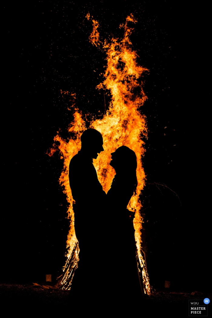 La mariée et le marié se tiennent devant un grand feu de joie la nuit sur cette photo prise par un photographe de mariage du New Jersey.