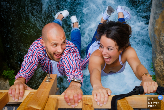 The bride and groom hang off of a bridge over white water in this photo by an Alicante, Valencia wedding photographer. 