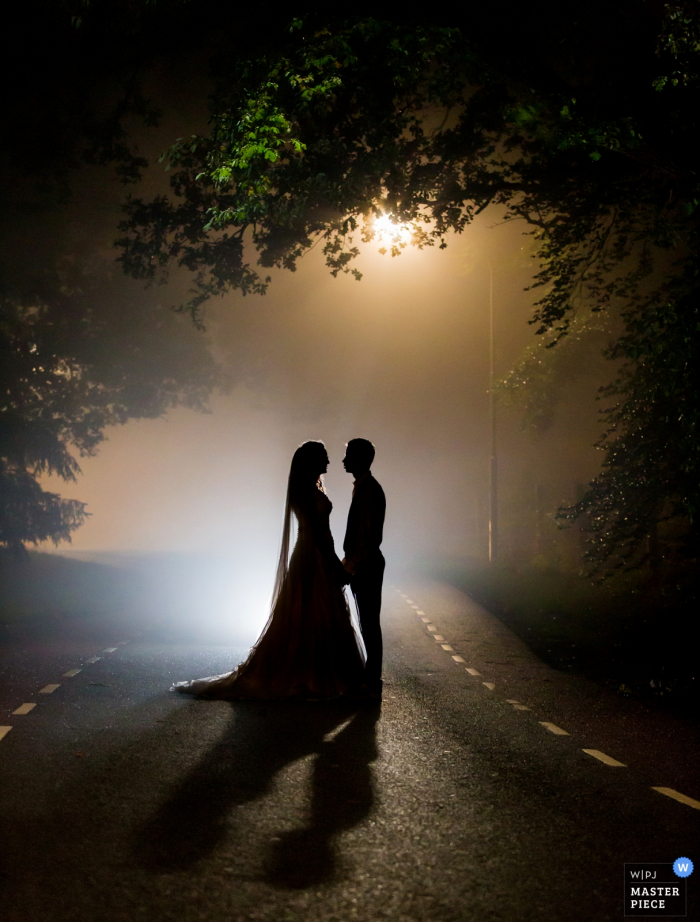 Foto der Braut und des Bräutigams, die durch eine Straßenlaterne silhouettiert werden, wie sie auf einer Straße nachts von einem niederländischen Hochzeitsfotografen stehen.