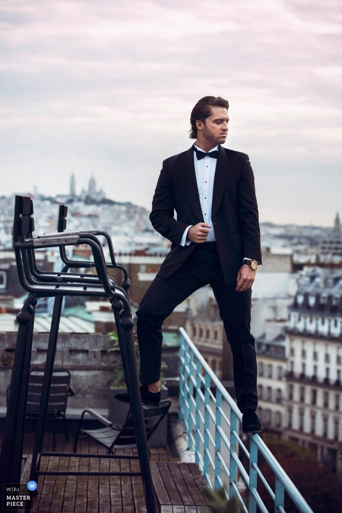Photo by: <a href="https://www.wpja.com/contests/2017-tri3-photography-competition/wildcard-portrait/2153351">Julien Scussel - France</a> Paris, France wedding day portrait of the groom standing on a chair and a roof top guard rail as he looks into the distance