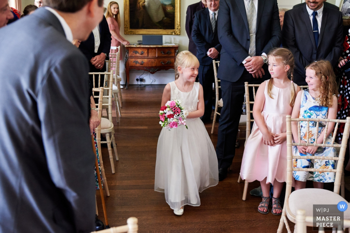 Foto von: <a href="https://www.wpja.com/contests/2017-tri3-photography-competition/kids-being-kids/2155476"> Ian Bursill - Großbritannien </a> Ein Hochzeitsfoto des Blumenmädchens aus Leicestershire, East Midlands, das den Gang entlang geht, während sie zwei kleine Mädchen ansieht