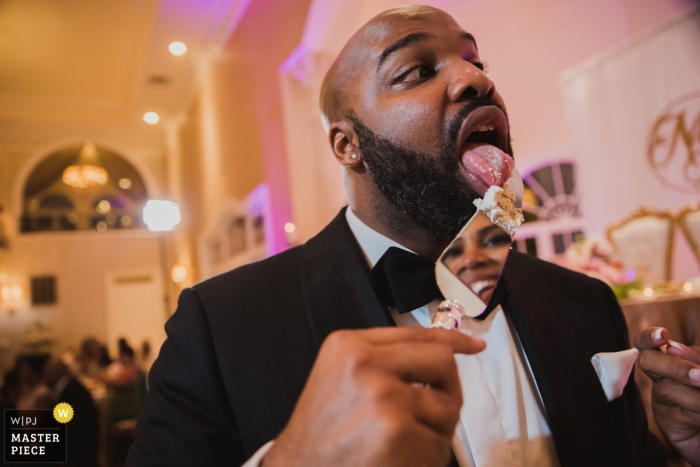 Photo by: <a href="https://www.wpja.com/contests/2017-tri3-photography-competition/cake-cutting/2107011">Ken Pak - United States</a> The bride's face is visible in the reflection of the cake server as the groom licks the server clean in this photo by a Washington, D.C. wedding photographer.