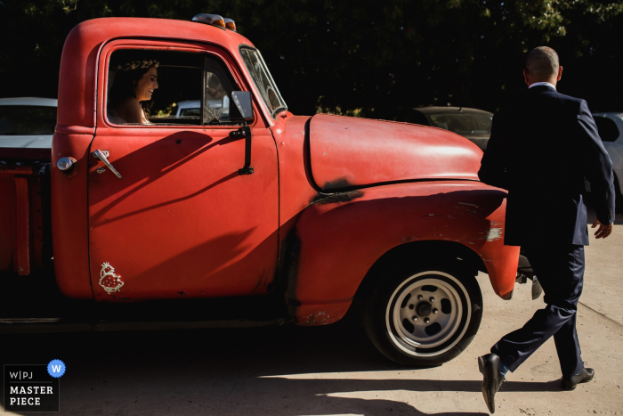 Lo sposo cammina intorno a un camion rosso per far uscire la sposa dall'altra parte in questa foto di un fotografo di matrimoni Cabo San Lucas.