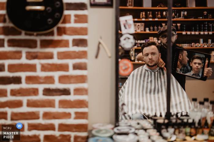 A barber holds up a mirror for the groom in this photo taken from outside in Centre-Val de Loire A barber holds up a mirror for the groom in this photo taken from outside by a Centre-Val de Loire wedding photographer.