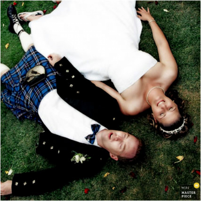 The bride and groom in his kilt are lying down on the grass in Manitoba