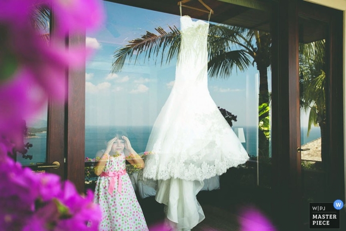 Il fotografo di matrimoni di Koh Samui ha catturato questa foto di una bambina che ammirava l'abito da sposa attraverso una grande vetrata