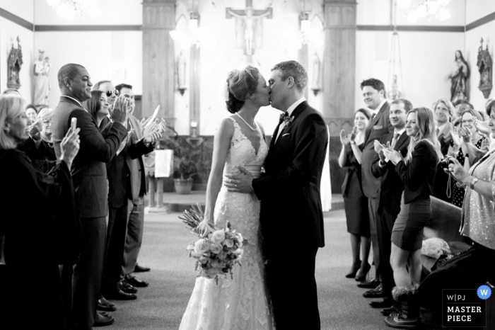 Um fotógrafo de casamento capturou esta foto em preto e branco dos noivos se beijando na cerimônia enquanto os convidados comemoravam