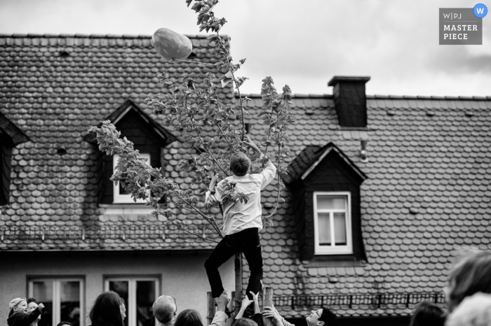 Der Bräutigam klettert in Hessen, Deutschland, auf einen Baum, um einen Ballon zu retten, während Hochzeitsgäste in einem unvergesslichen Schwarz-Weiß-Außenbild zuschauen