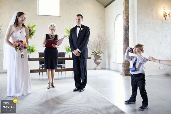 Un giovane fotografo in erba viene catturato mentre gli sposi di Melbourne sorridono al loro servizio di nozze all'interno di una chiesa in Australia