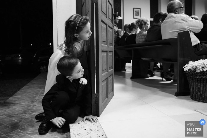 Photographer captures two very young wedding guests peeking through the doorway to observe an indoor Santa Catarina wedding ceremony in Brazil