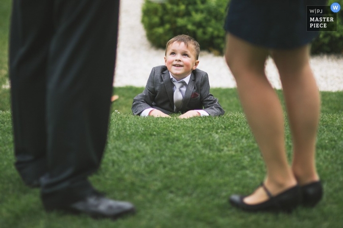 Image of a young suited lad lying on the grass looking up at his wedding guest parents while they celebrate in an outdoor setting