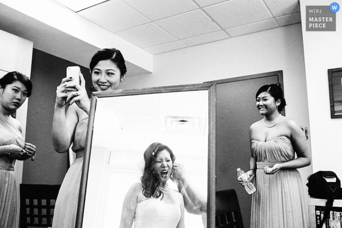 A black and white photo of a Bronx bride being assisted with the final jewelry for her wedding look reflected in a full length mirror surrounded and supported by her bridesmaids in NY