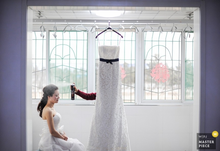 Laca para el cabello frente a una ventana para una novia de Shanxi mientras se prepara para vestirse con una fotografía deliberada de China con líneas severas suavizadas por el vestido que cuelga esperando a la novia