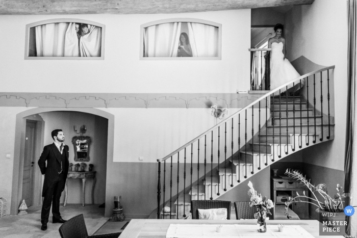 Aquí viene la novia mientras baja hasta el final de las escaleras donde su novio la espera en una fotografía de boda en interiores en blanco y negro de Morbihan Brittany.