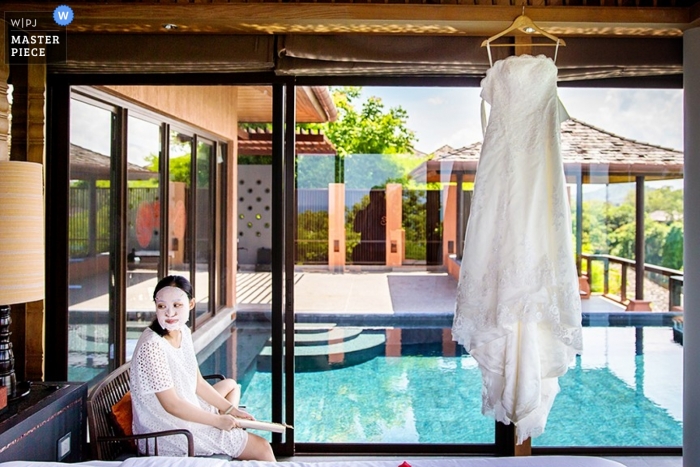 Una sposa di Phuket aspetta di vestirsi per il suo matrimonio con il suo abito appeso alla porta di un'area di una piscina blu brillante che crea la scena per una bella immagine del matrimonio di destinazione Thailandia