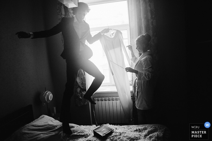 With a bounce on the bed a St Petersburg groom hands his bride her wedding dress while winter sunlight streams into a bedroom in Russia