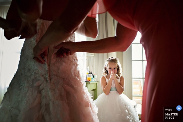 Foto von: <a href="https://www.wpja.com/contests/2014-q4-photography-contest/getting-ready/1555881"> Julie Ambos - Vereinigte Staaten </a> Der Hochzeitsfotograf von Key West macht ein Bild von einem überraschten Gesicht eines kleinen Mädchens, als sie zusieht, wie das Hochzeitskleid auf die Braut geschnürt wird