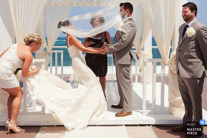 A bridesmaid helps the bride with her train as her veil blows into the groom's face in this documentary-style photo by a Playa del Carmen photographer.