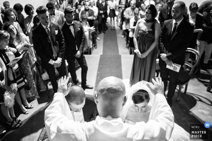 The priest holds his hands over the bride and groom as they kneel at the altar in this black and white photo taken by a documentary-style Tuscany wedding photographer.