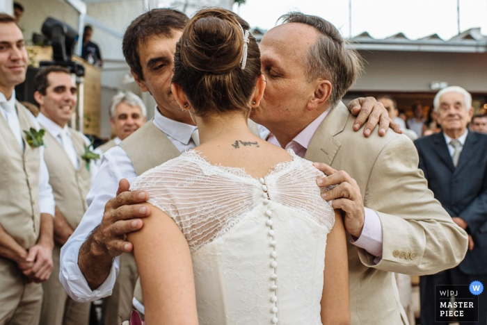 Rio de Janeiro wedding photographer captured this photo of the bride being kissed on the cheek by her father as the rest of the wedding guests watch nearby