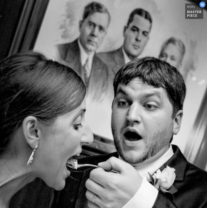 Le photographe de mariage de Baton Rouge a capturé cette photo du marié qui nourrissait la mariée avec un morceau de gâteau devant un tableau représentant des hommes avec des regards désapprobateurs