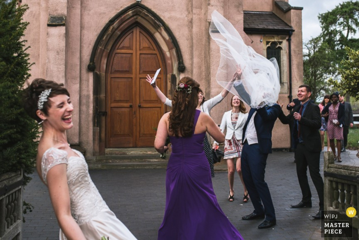 Festa nupcial em Londres rindo após a cerimônia de casamento enquanto o véu da noiva voa pelo ar - Fotojornalismo de reportagem de casamento na Inglaterra