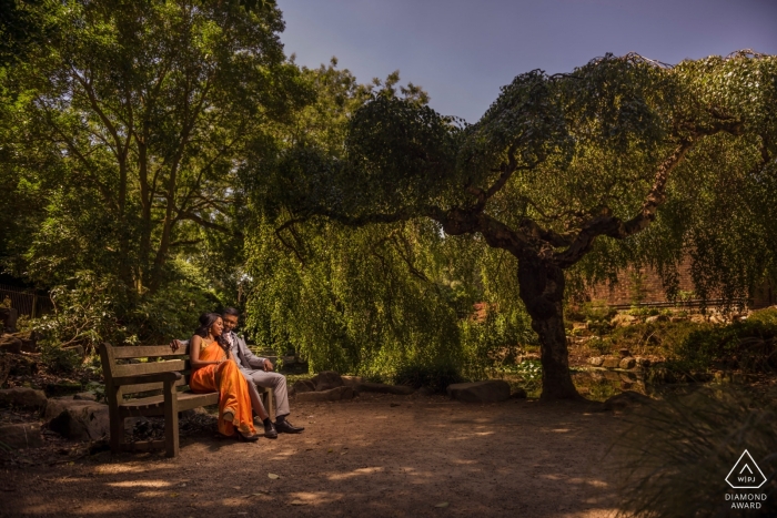 Fotos de noivado na Inglaterra de um casal sentado em um banco com bela sombra de árvore | Sessão pré-casamento de fotógrafo no Reino Unido com fotógrafo