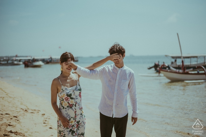 Indonesia engagement pictures of a couple covering each others eyes at the beach with boats  | Bali photographer pre-wedding shoot with photographer