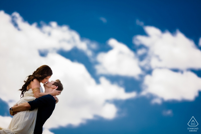 Foto: <a href="https://www.wpja.com/photos/diamond-award-top-wedding-engagement-photographer/2338371"> Joshua Grasso - Buford, Georgia Georgia Hochzeit Verlobungsportrait eines Paares, das unter dem großen blauen Himmel mit Wolken küsst Atlanta-Hochzeit vor der Hochzeit