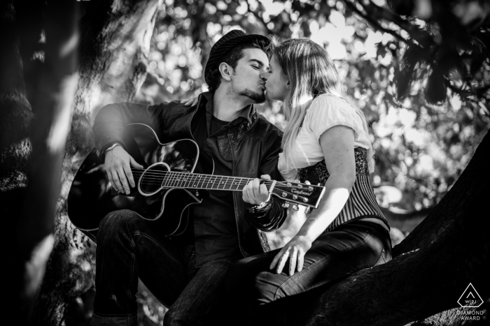 Angleterre Photos noir et blanc de fiançailles d'un couple jouant de la guitare dans les arbres en s'embrassant | Séance de portrait pré-mariage d'un photographe britannique