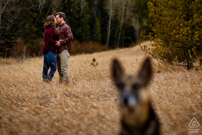 Das verlobte Paar aus Denver, CO, küsst sich in der Nähe von Krileys Teich, während ihr Hund Koda während der Verlobungsfotografie im Frühling im Golden Gate Canyon Ausschau hält.