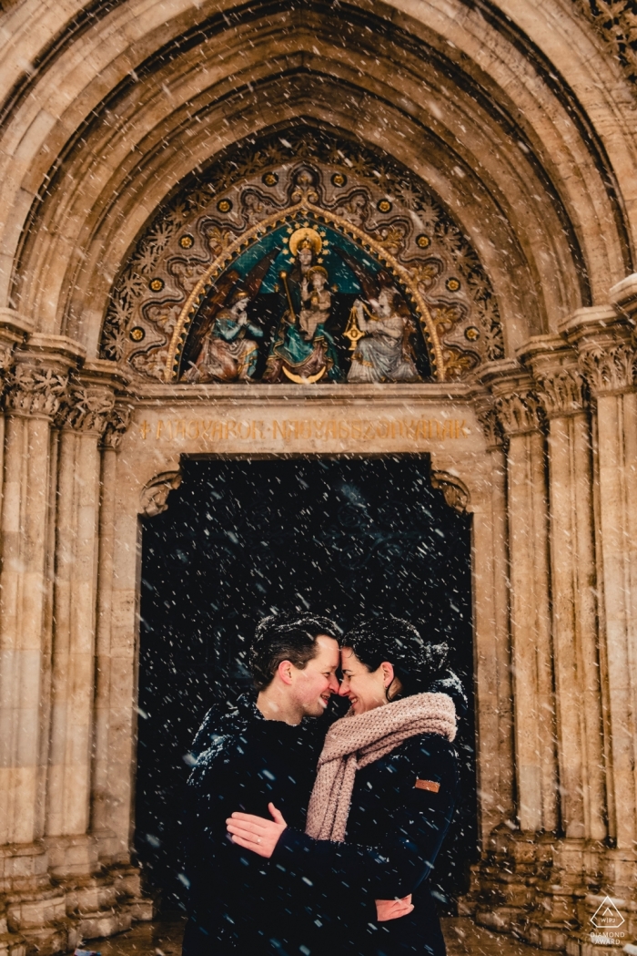 Budapest couple stands in a snowstorm, framed by an arched doorway to a majestic cathedral