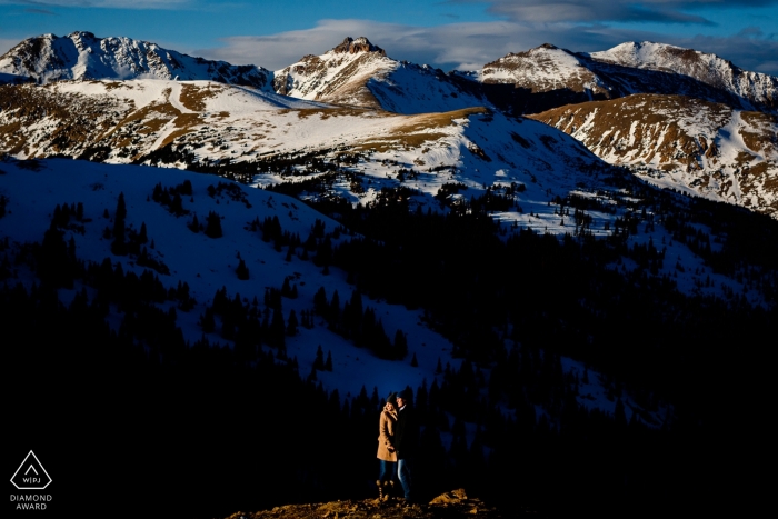 The Colorado engaged couple is standing above a mountain range covered in snow