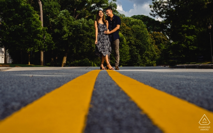 In Atlanta, Georgia, a playful engaged couple smiles for the camera while standing on bold yellow traffic lines in the roadway, viewed from a low angle