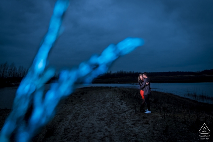 An engaged couple stands illuminated in a moment of embrace on a desolate beach in Utrecht, surrounded by a blue dusky palette