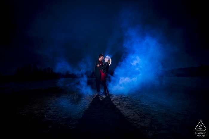 Dans cette scène éthérée, un couple nouvellement fiancé se tient sur une plage lisse et rocheuse à Utrecht, illuminée par le clair de lune et embrassée par le brouillard bleu remplissant le ciel nocturne