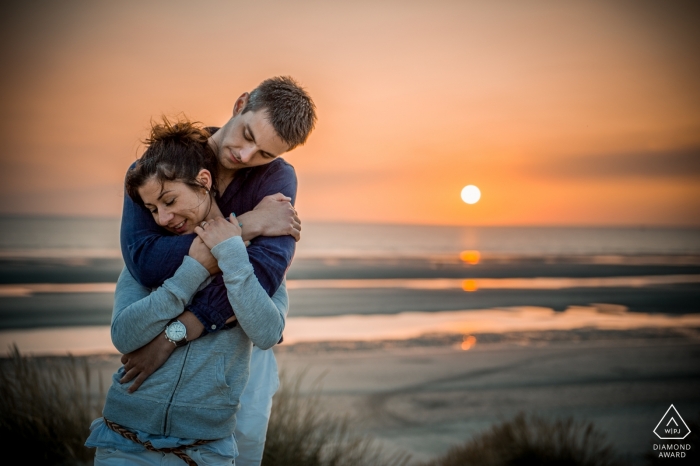Photo by: <a href="https://www.wpja.com/photos/diamond-award-top-wedding-engagement-photographer/2428506">Cedric Derbaise - Bury A young beautiful engaged couple embrace in a hug with their backs to the setting sun and the ocean