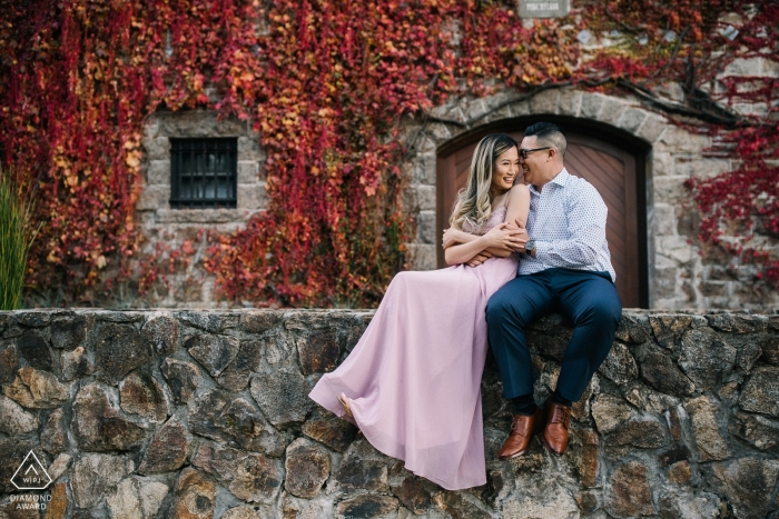 Sesión de retratos de compromiso de una pareja sentada junto a un muro de piedra frente a un edificio cubierto de viñedos en San Francisco.