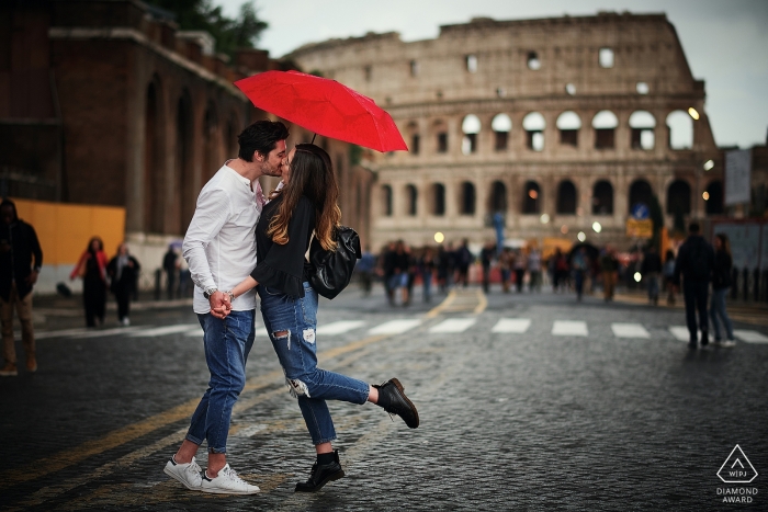 Un amorevole fidanzamento ritratto di una coppia che si bacia sotto un ombrello rosso di fronte al Colosseo a Roma, Italia.