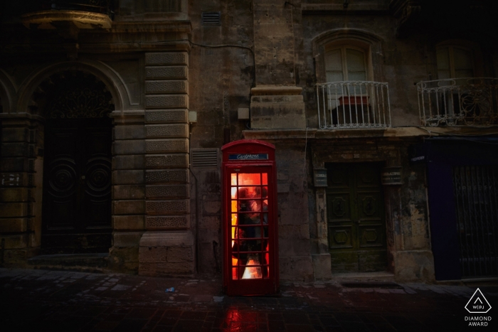 Engagement Portrait Session showing a couple snuggles up in a red phone box at night Valleta Malta Engagement Portrait Session - A couple snuggles up in a red phone box at night in Malta
