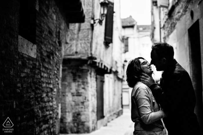 Photo by: <a href="https://www.wpja.com/photos/diamond-award-top-wedding-engagement-photographer/2457016">Franck Petit - Serignac sur Garonne Black and white engagement photo of a couple about to kiss in a narrow street surrounded by old stone buildings in Agen, France.