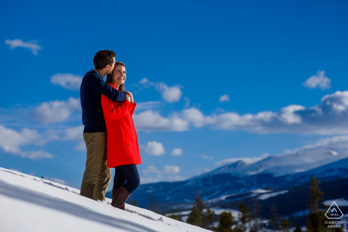 Photo by: <a href="https://www.wpja.com/photos/diamond-award-top-wedding-engagement-photographer/2457586">Justin Edmonds - Denver Colorado Sapphire Point Engagement Photography Session - Kisses in the snow wearing a read winter coat as Breckenridge resort is seen in the background