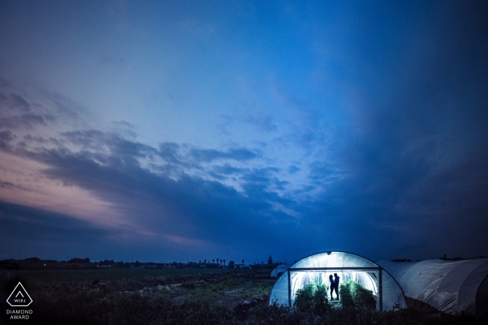 Engagement photo session of a couple standing in a lit up greenhouse at dusk in Siracusa.