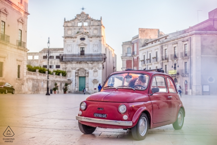 Engagement photo of a couple driving in a small red car through Siracusa. 